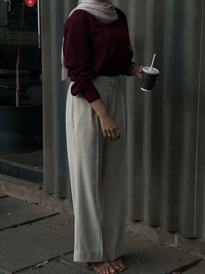 Woman in maroon long sleeve top and cream wide-leg trousers holding a drink.