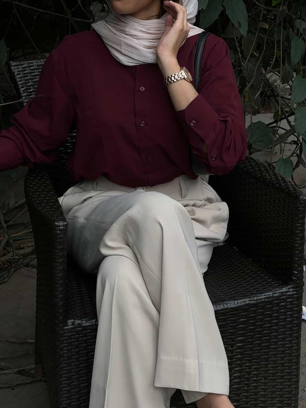 Woman wearing a maroon button-down shirt and beige wide-leg trousers sitting in a wicker chair.