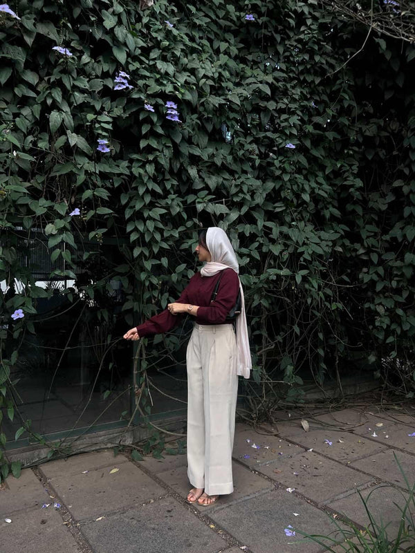 Woman in maroon long sleeve top and cream wide-leg trousers posing against a green vine wall.