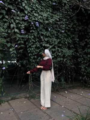 Woman in maroon long sleeve top and cream wide-leg trousers posing by a green leaf wall.