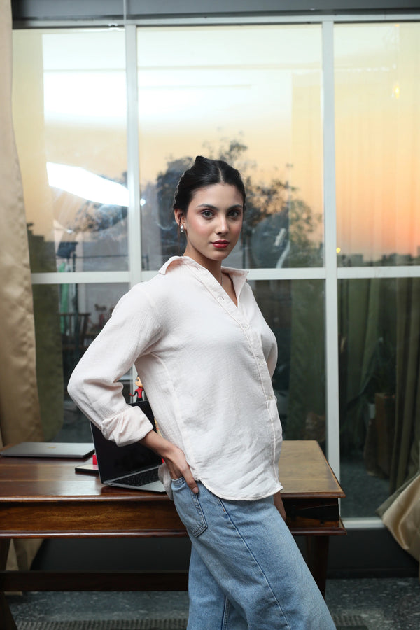 Woman in white button-down shirt and blue jeans posing by a window at sunset.