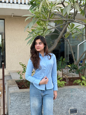 Young woman in a light blue button-down shirt and denim jeans standing by a tree.