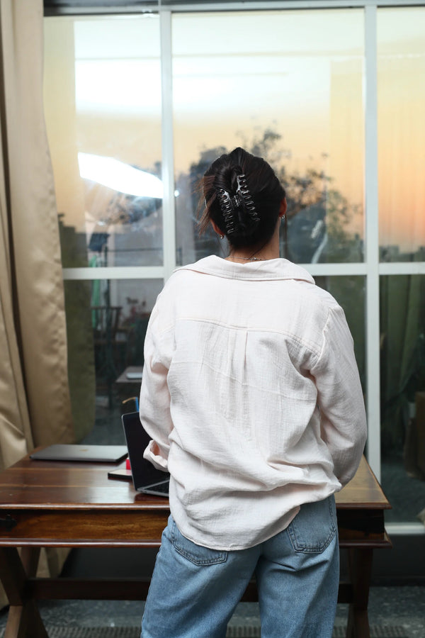 Woman in a white linen shirt and blue jeans standing by a window at sunset.