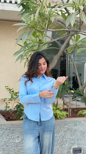 Woman in a light blue button-down shirt and baggy denim jeans standing by a tree.