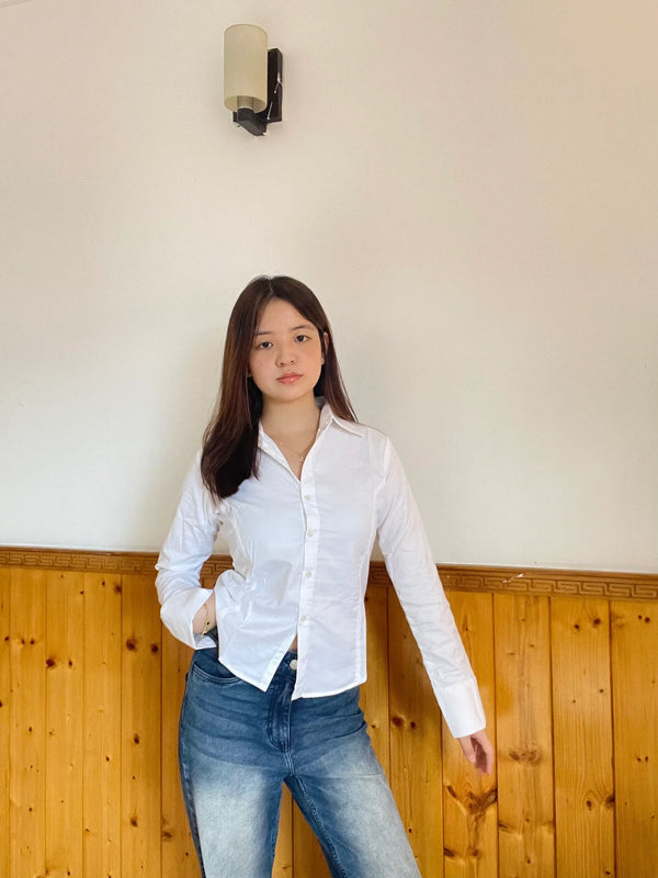 Woman wearing a white button-down shirt and blue denim jeans standing against a wooden wall.