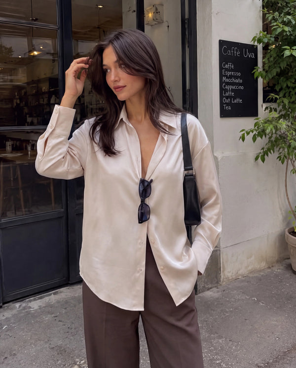 Woman in a cream satin long sleeve shirt and brown trousers standing by a cafe.
