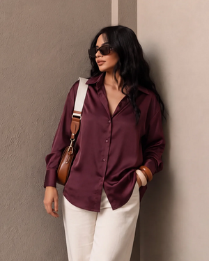 Woman wearing a maroon satin button-down shirt, white trousers, and sunglasses with a brown bag.