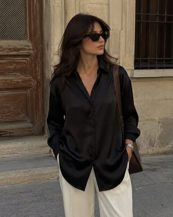 Woman wearing a black satin button-down shirt and white trousers with sunglasses.