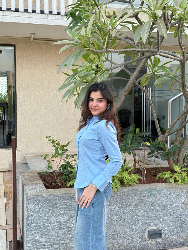Young woman wearing a light blue button-down shirt and denim skirt posing by a tree.
