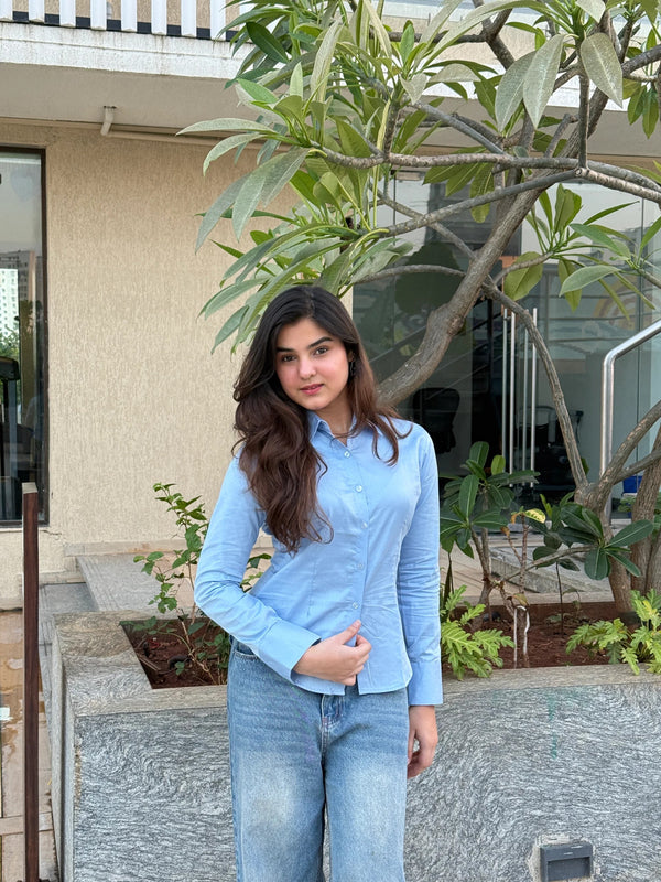 Woman wearing a light blue button-down shirt and denim jeans standing by a tree.